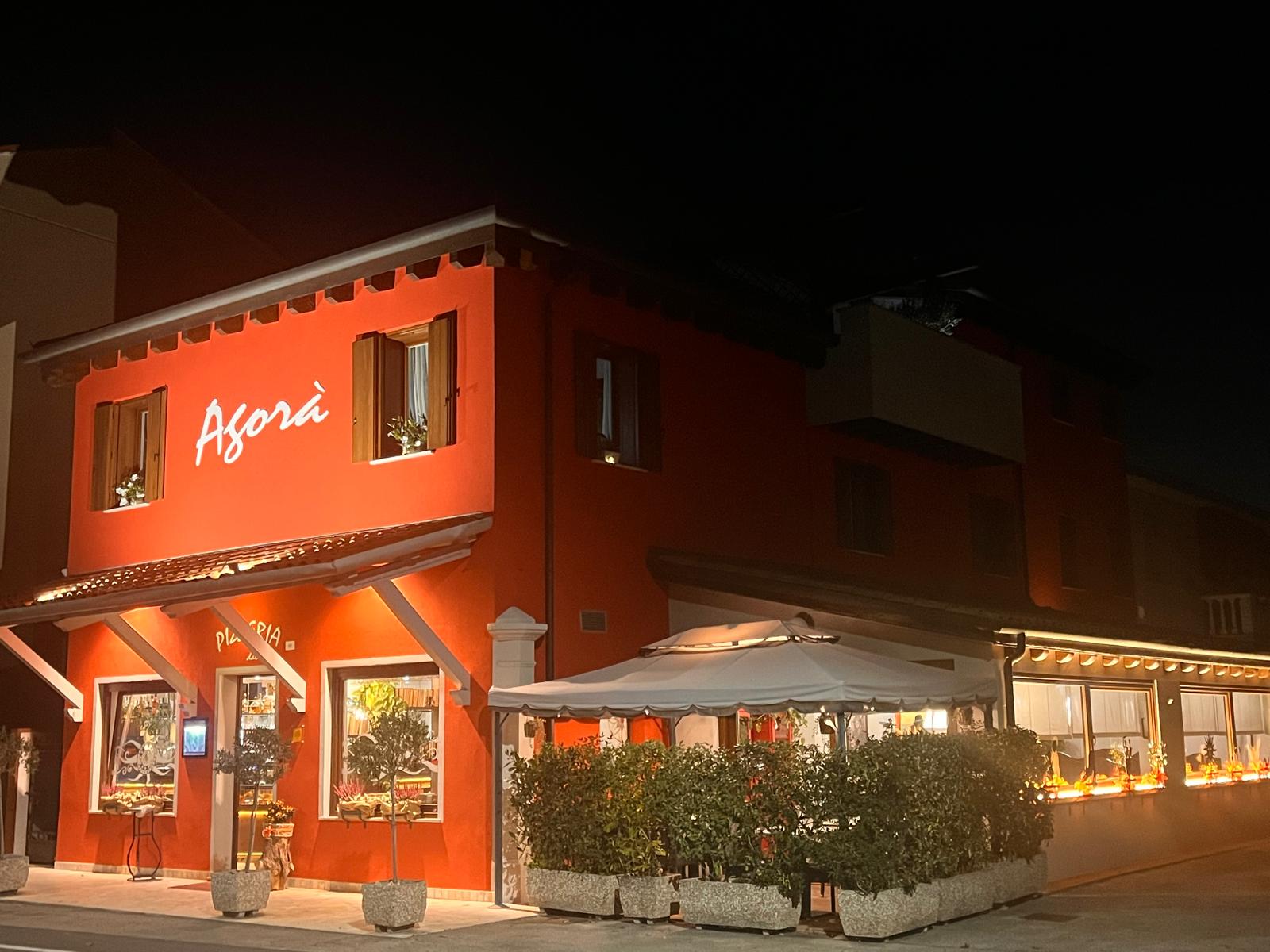 La facciata di Pizzeria Agorà illuminata di sera, con l’insegna luminosa e gli ulivi in vaso davanti all’ingresso